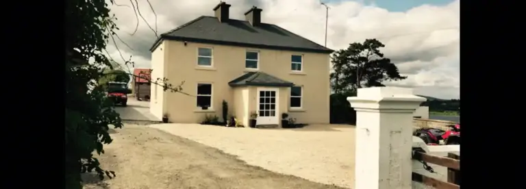 A cream-coloured two-storey house with a grey roof, large front windows, and a small entrance porch, set in a rural area with a gravel drive and countryside in the background.