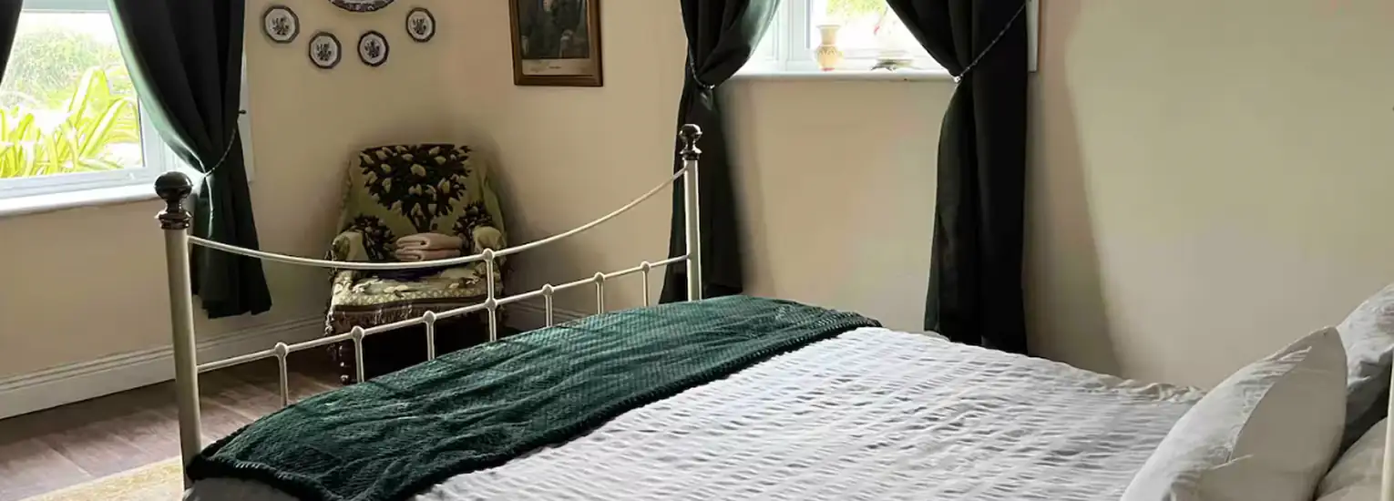 A cosy bedroom with a metal bed frame, white bedding, and a dark green throw blanket. There are two windows with dark curtains, an armchair in the corner, and decorative plates on the wall above the chair.