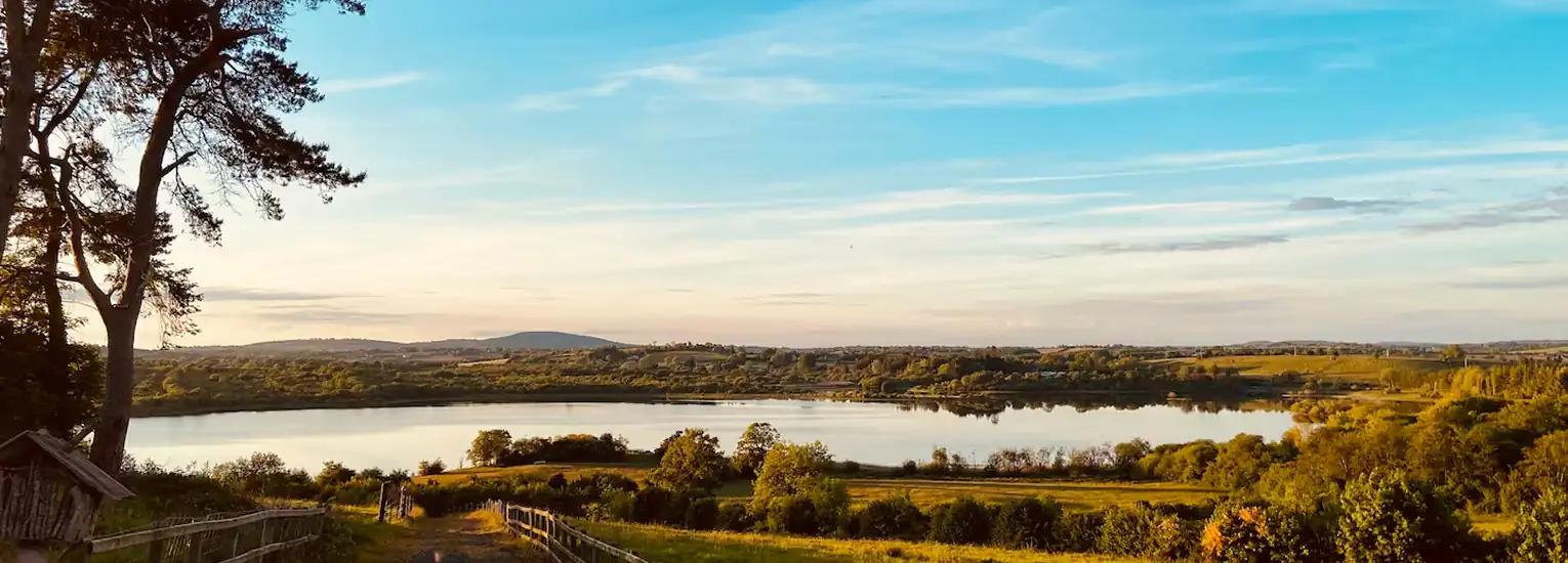 A scenic view of a calm lake surrounded by green fields and trees under a blue sky with light clouds, taken at sunset with warm golden light. A wooden fence runs along a path in the foreground.