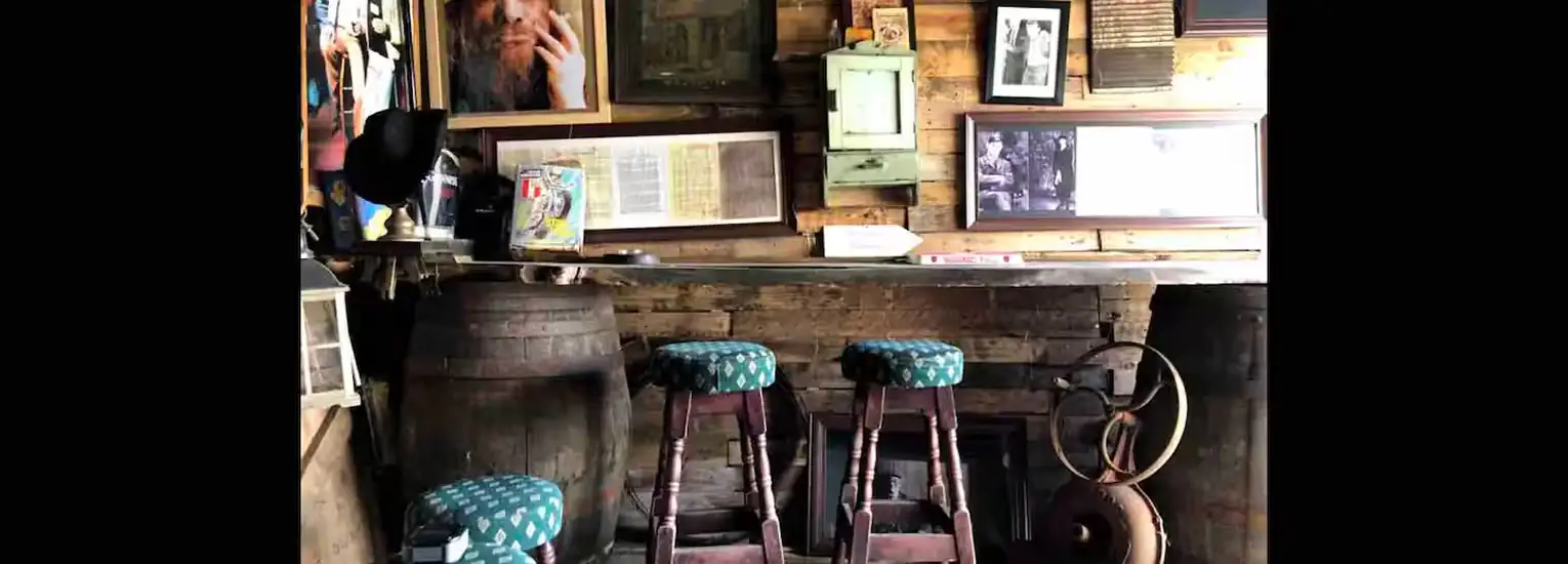 A rustic bar interior with three wooden stools, each with a blue patterned seat, stands in front of a wooden counter. Barrels, vintage photos, books, and framed portraits decorate the walls and shelves.