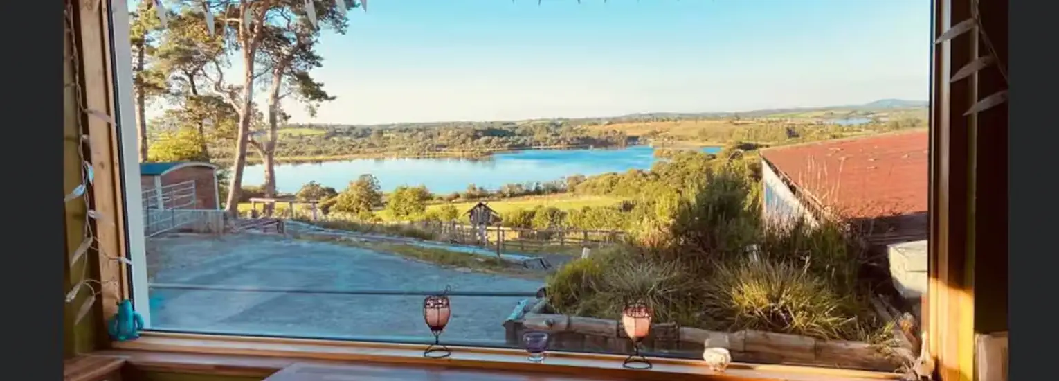 A large window offers a scenic view of a lake surrounded by green fields, trees, and distant hills under a clear blue sky. A wooden table and decorative candle holders are visible by the window.