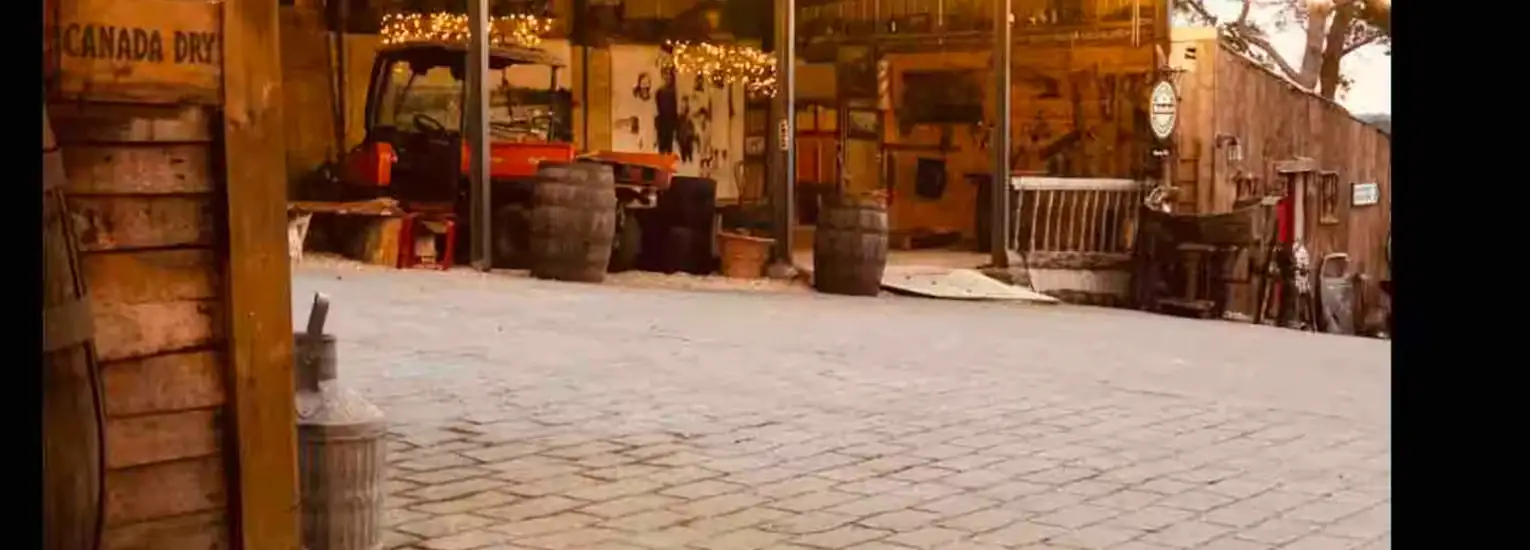 A rustic outdoor scene featuring a brick-paved area, wooden barrels, various antique items, and a partially visible orange tractor under warm fairy lights near a wooden building.