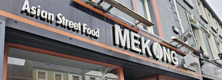 A restaurant sign reading "Asian Street Food MEKONG" is shown on the exterior of a building, with part of the street and other buildings reflected in the windows below.