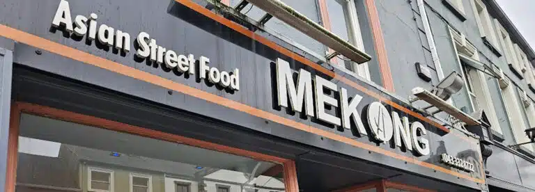 A restaurant sign reading "Asian Street Food MEKONG" is shown on the exterior of a building, with part of the street and other buildings reflected in the windows below.