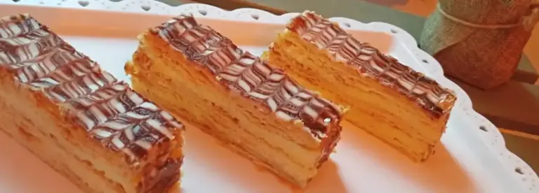 Three rectangular slices of layered pastry with a white and chocolate zigzag icing pattern on top, arranged neatly on a white decorative tray.