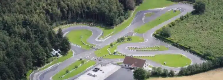 Aerial view of a go-kart racing track with winding asphalt lanes, bordered by green grass and trees. A small building with a car park is visible near the track’s entrance.