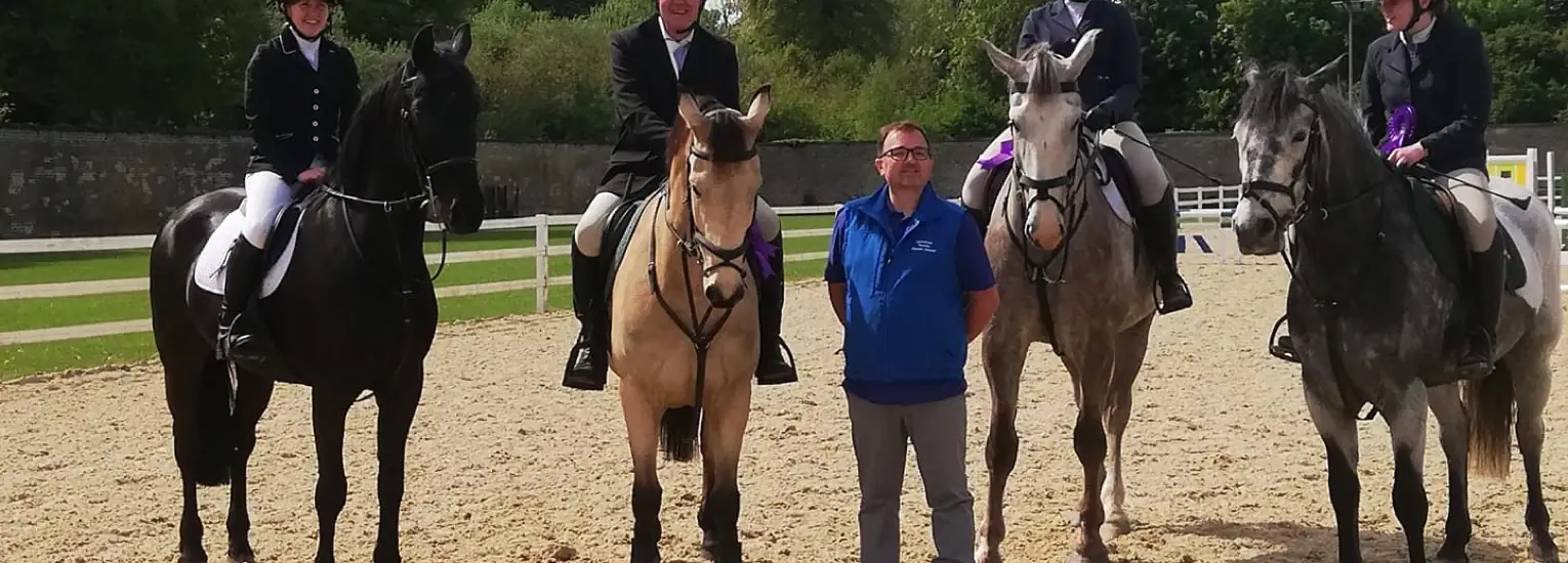 Four people in equestrian attire sit on horses in a sandy arena, whilst a man in a blue shirt stands between two horses. Trees and a fence are visible in the background.