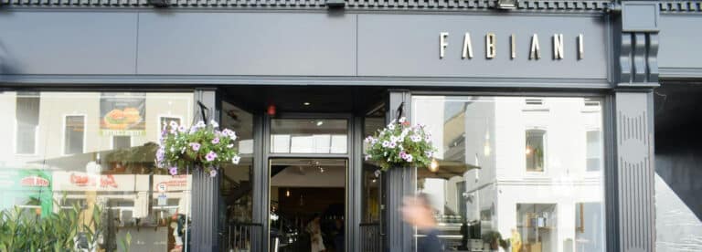 Street view of the entrance to Fabiani, a modern shopfront with large glass windows, black exterior, hanging flower baskets, and the Fabiani sign above the door. A person is walking past the entrance.
