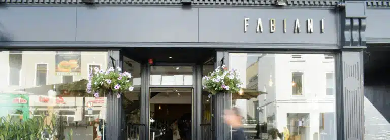 Street view of the entrance to Fabiani, a modern shopfront with large glass windows, black exterior, hanging flower baskets, and the Fabiani sign above the door. A person is walking past the entrance.