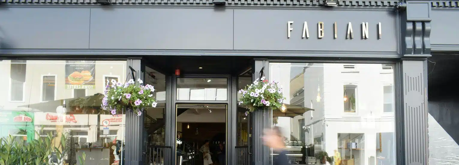 Street view of the entrance to Fabiani, a modern shopfront with large glass windows, black exterior, hanging flower baskets, and the Fabiani sign above the door. A person is walking past the entrance.