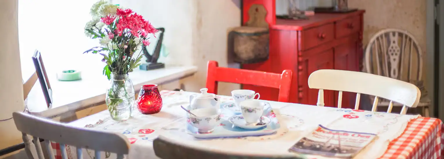 A cosy dining area with a table set for tea, featuring floral-patterned teacups, a bouquet of flowers in a jar, a red candle holder, and a newspaper, with mismatched chairs and a red side cupboard in the background.