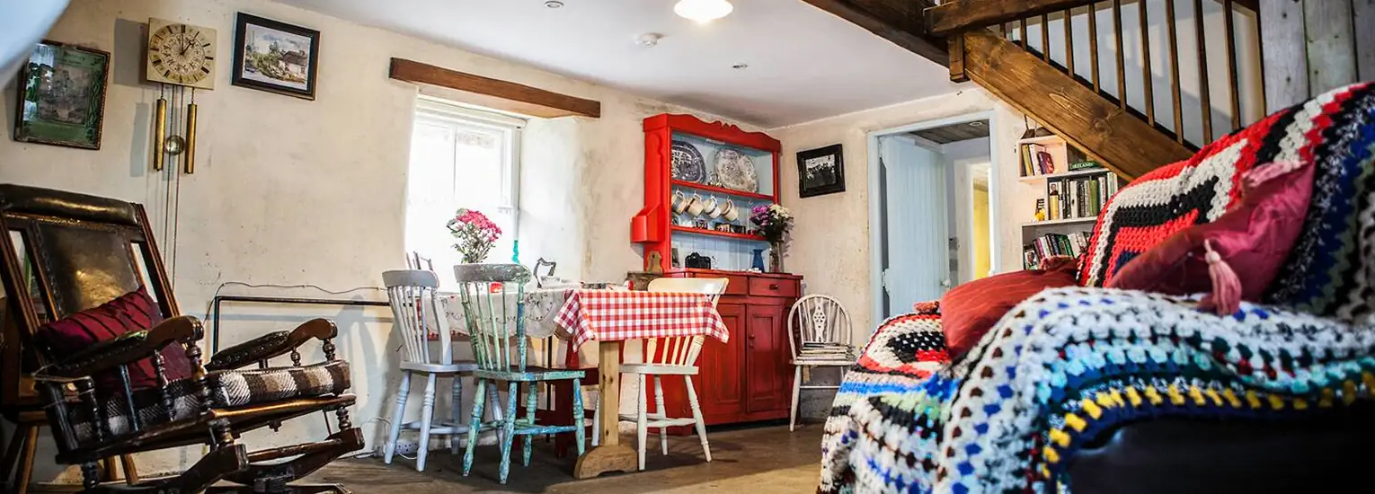 A cosy rustic living room with mismatched chairs around a table with a red checked tablecloth, a red cupboard, a crocheted blanket on a sofa, a rocking chair, and wooden stairs. Sunlight shines through a window.