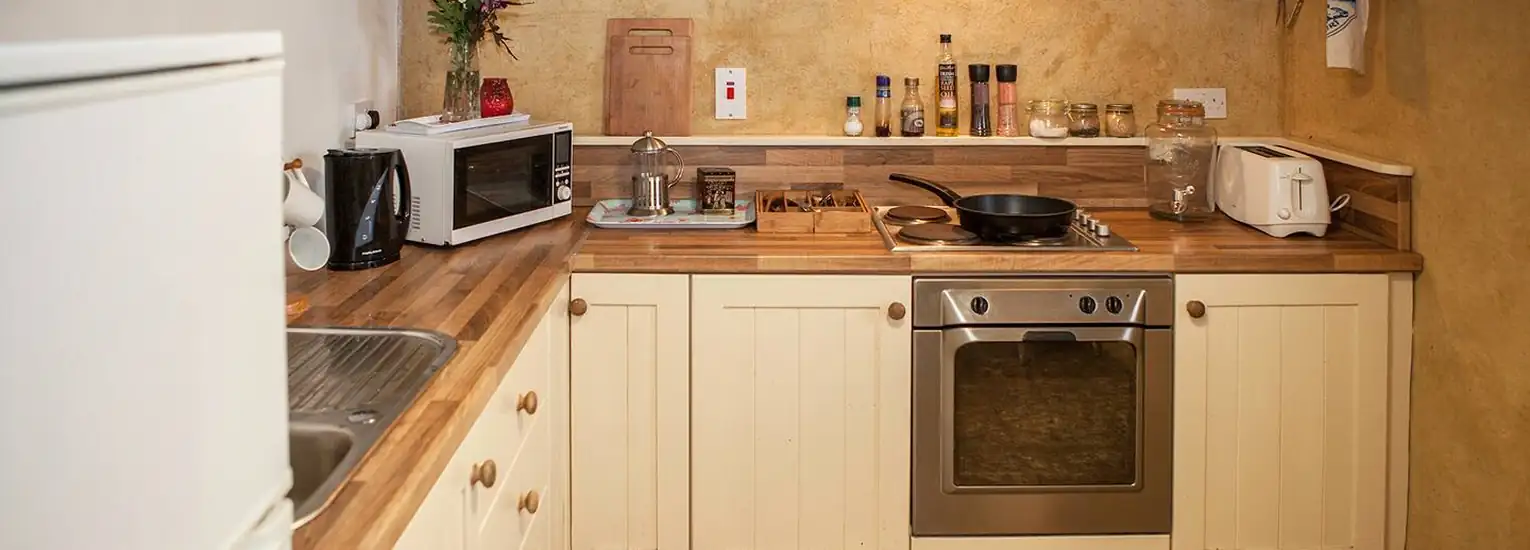 A cosy kitchen with cream cupboards, wooden worktops, an oven with a hob, microwave, toaster, kettle, jars, and various utensils neatly arranged against a warm yellow wall.