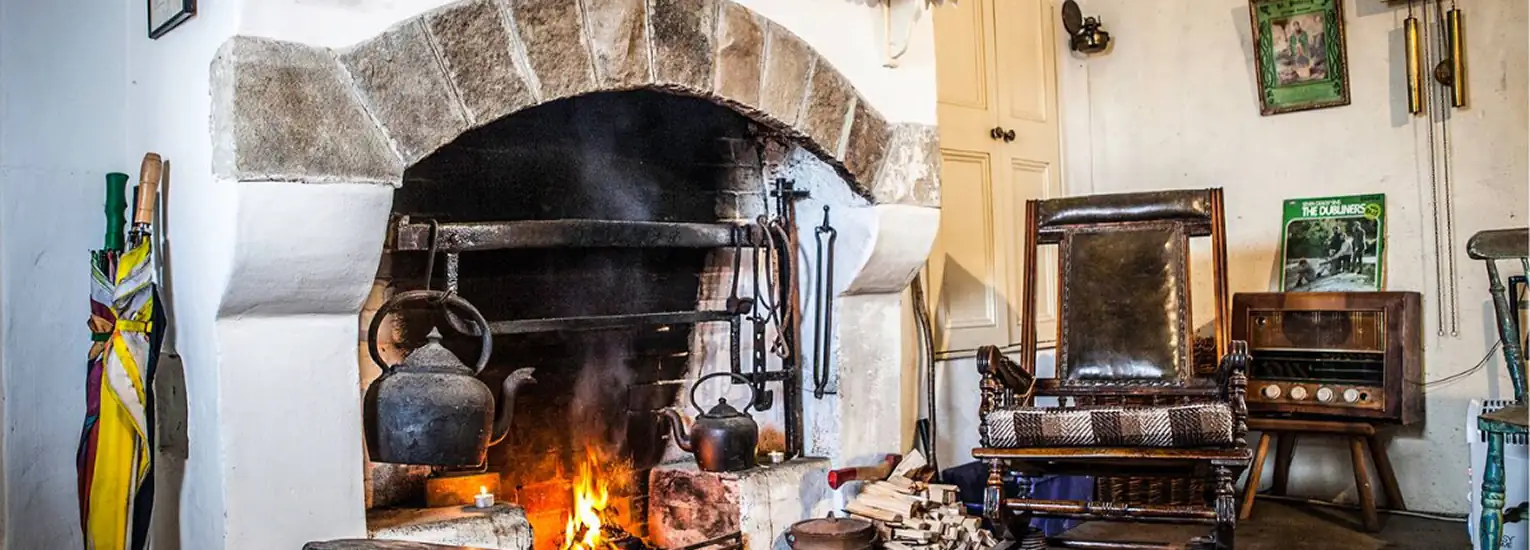 A cosy room with a stone fireplace, iron kettles hanging above a fire, rocking chair, stacked firewood, vintage radio, and rustic decor, including umbrellas and framed pictures on the wall.