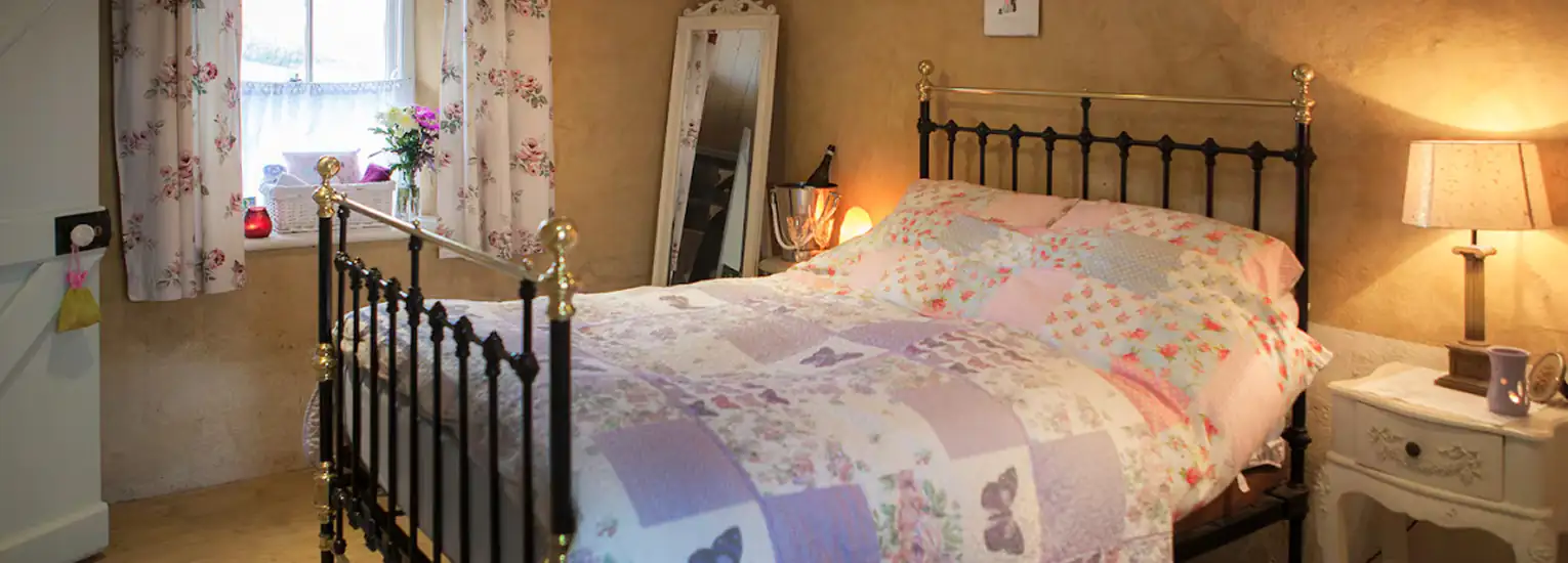 A cosy bedroom with a black metal bed frame, patchwork quilt, bedside table with lamp, flowers on the windowsill, a standing mirror, and soft warm lighting. The walls are beige and rustic.