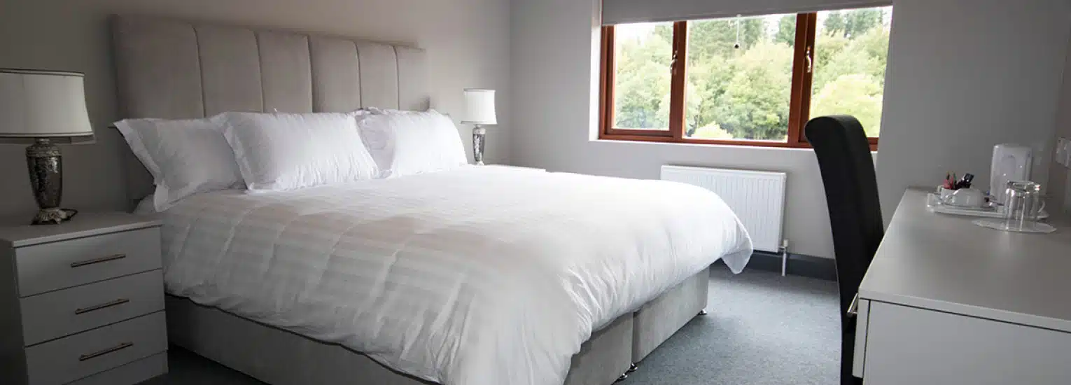 A bright, modern bedroom with a large bed covered in white linen, two bedside tables with lamps, a desk with a chair, and a window overlooking greenery. The decor is minimalist and clean.