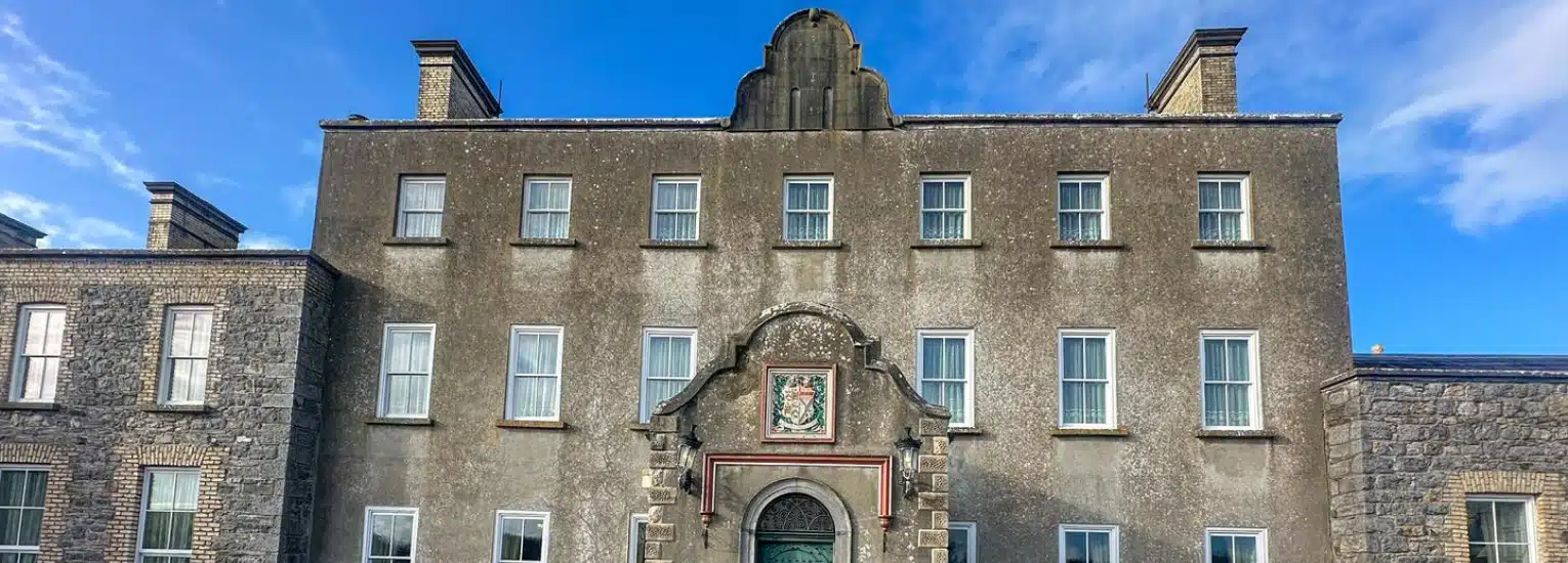 A large historic stone building with symmetrical windows, a decorative arched entrance, and a crest above the door, set against a blue sky with scattered clouds.
