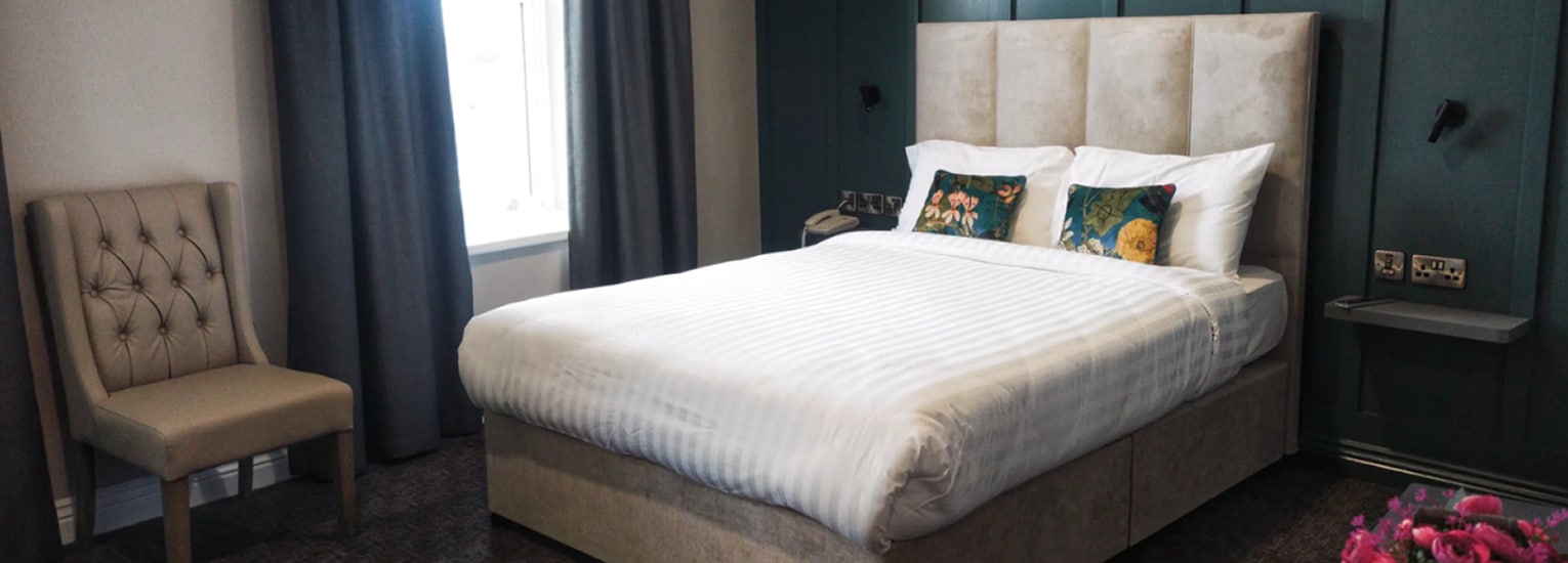 A modern hotel room with a large bed featuring white linen and floral cushions, a tufted beige chair, dark green feature wall, grey curtains, and a window letting in natural light.