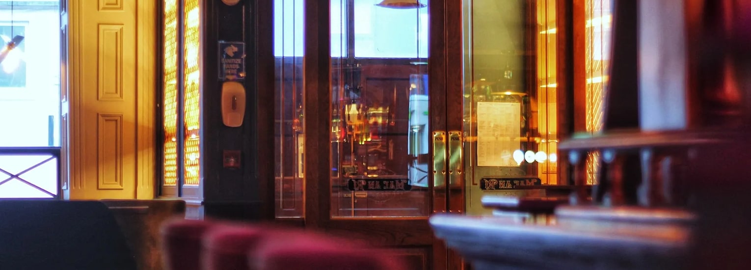 Warmly lit bar interior with wooden furniture, glass doors, and reflections of bottles. Soft yellow and orange lighting creates a cosy, inviting atmosphere. Some blurred elements in the foreground.