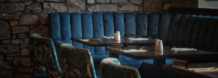 A dimly lit restaurant booth with blue velvet seating and stone wall background. Tables are set with menus, salt and pepper pots, cutlery, and upholstered chairs facing the booth.