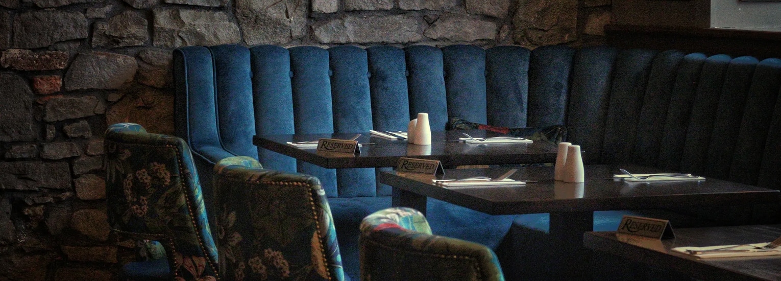 A dimly lit restaurant booth with blue velvet seating and stone wall background. Tables are set with menus, salt and pepper pots, cutlery, and upholstered chairs facing the booth.