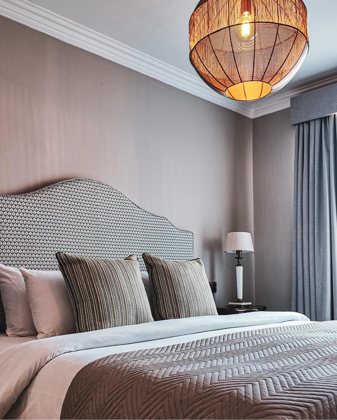 A modern bedroom with a patterned headboard, two large pillows, a textured duvet, a bedside table with a lamp, grey curtains, and a woven pendant light hanging from the ceiling.