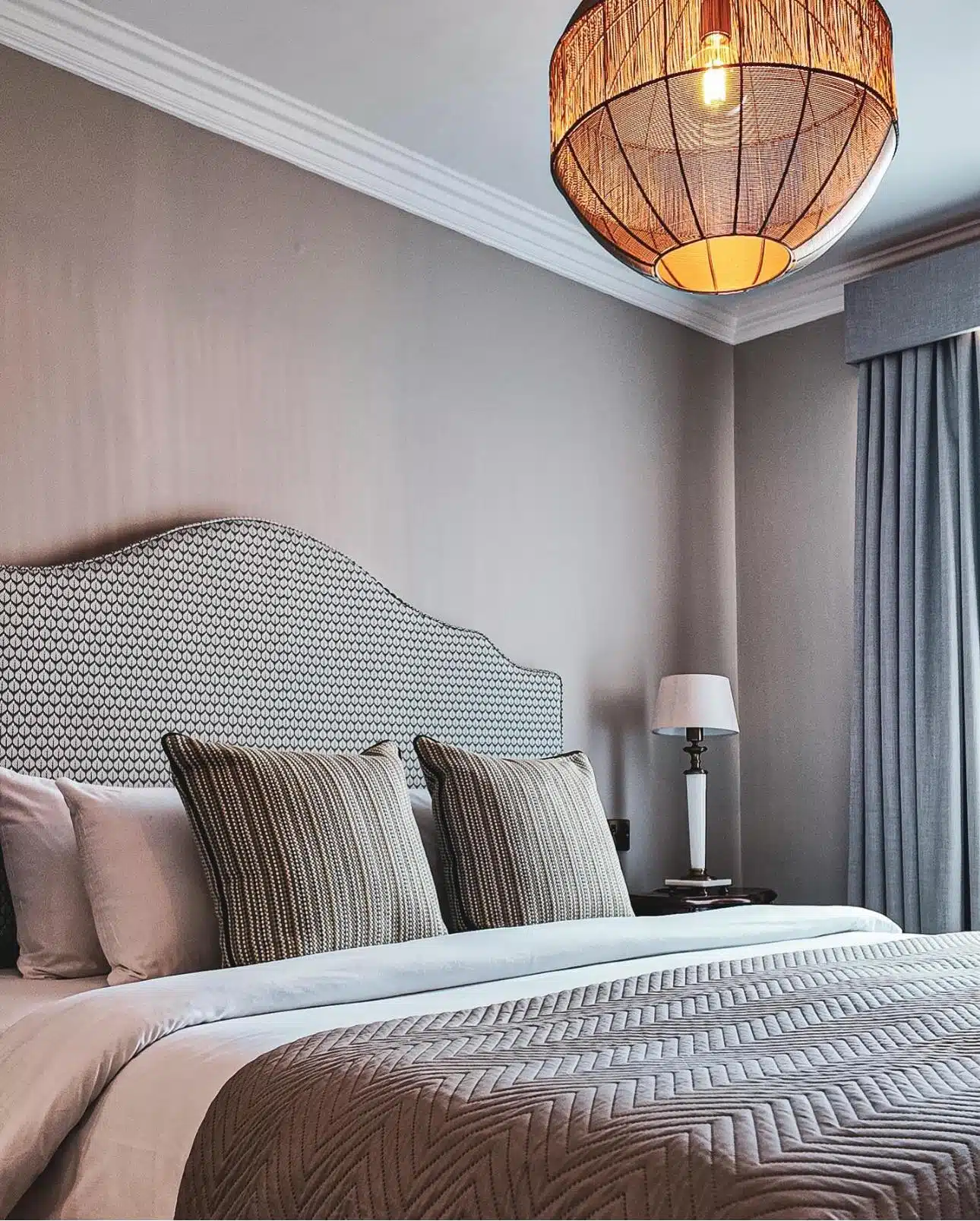A modern bedroom with a patterned headboard, two large pillows, a textured duvet, a bedside table with a lamp, grey curtains, and a woven pendant light hanging from the ceiling.