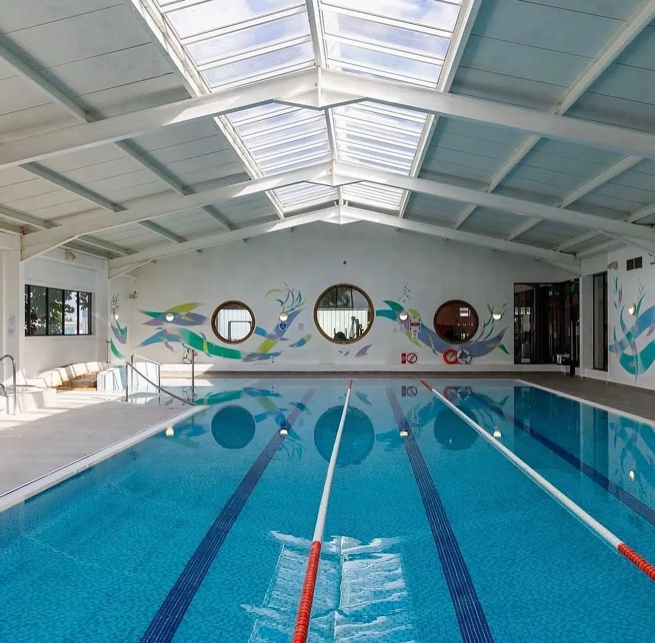 Indoor swimming pool with two swim lanes, blue tiled floor, and skylight ceiling. Wall features colourful fish murals and round windows. Sunlight shines through the glass roof, illuminating the bright, clean space.