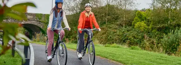 Two women wearing helmets and jackets ride bicycles on a paved path through a green park, with trees and a stone bridge in the background. Both women are smiling and enjoying the outdoor activity.