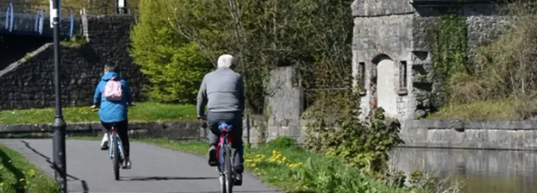 Two people ride bicycles along a paved path beside a canal with stone walls, greenery, and an old stone structure on the right. The scene is outdoors on a sunny day.