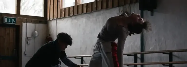 Two people in a dimly lit room; one person is sitting or kneeling and appears to be assisting or observing, whilst the other bends backwards dramatically during what seems to be a dance or movement practice.