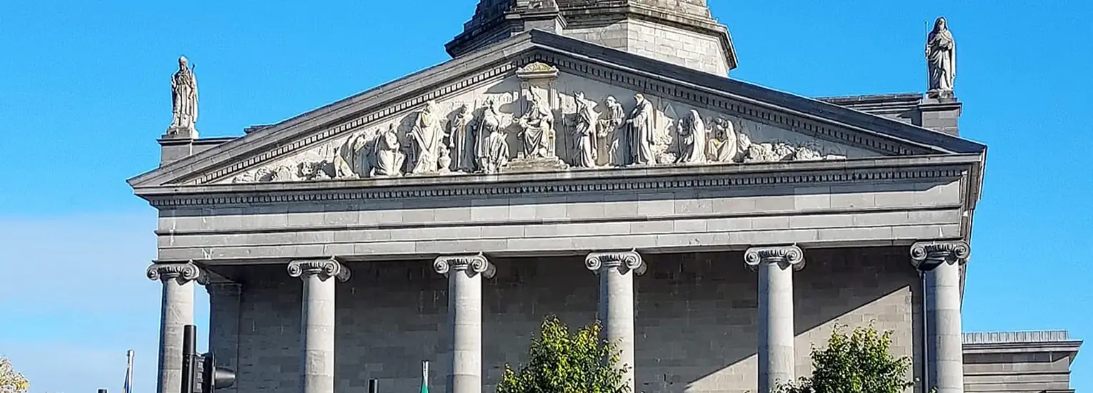 Neoclassical stone building with six large columns, triangular pediment adorned with sculpted figures, and statues at each end of the roof, set against a clear blue sky.