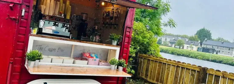 A red coffee van with an open service hatch displays cups, coffee machines, plants, and snacks. The van is parked near a wooden fence, overlooking a river and houses under a cloudy sky.