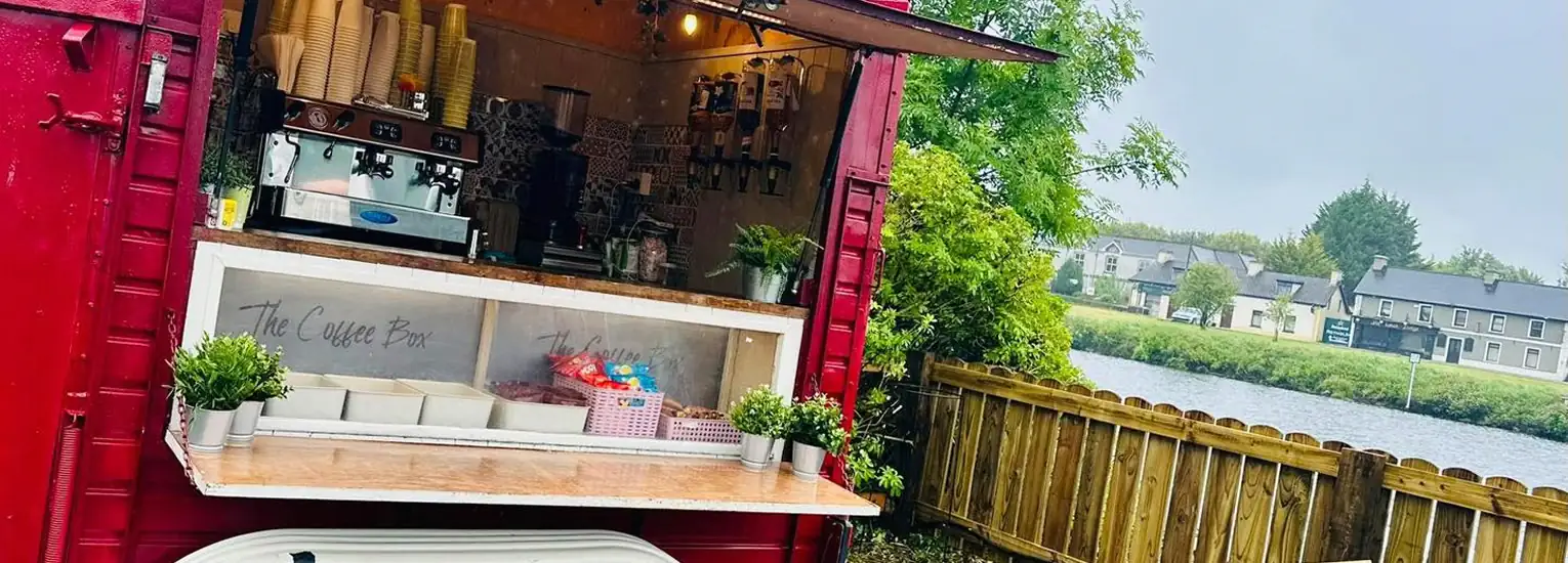 A red coffee van with an open service hatch displays cups, coffee machines, plants, and snacks. The van is parked near a wooden fence, overlooking a river and houses under a cloudy sky.