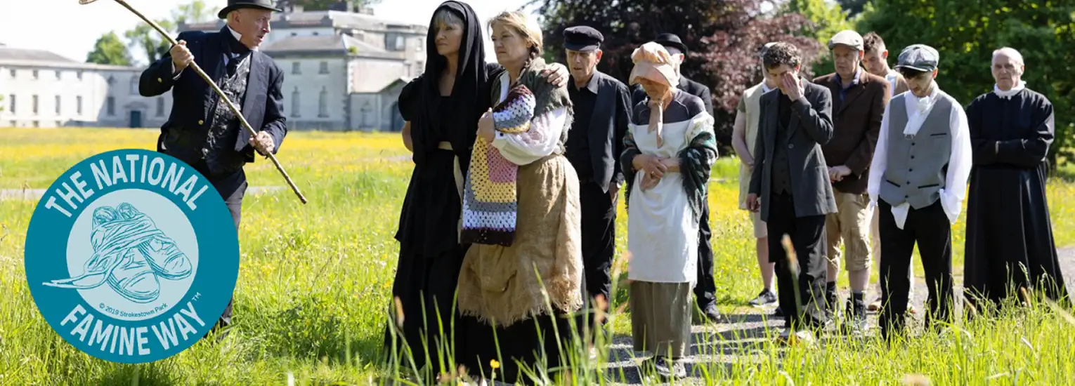 A group of people in historical clothing walk sombrely through a grassy field, led by a man with a staff. The National Famine Way logo is on the left. Trees and a large building appear in the background.
