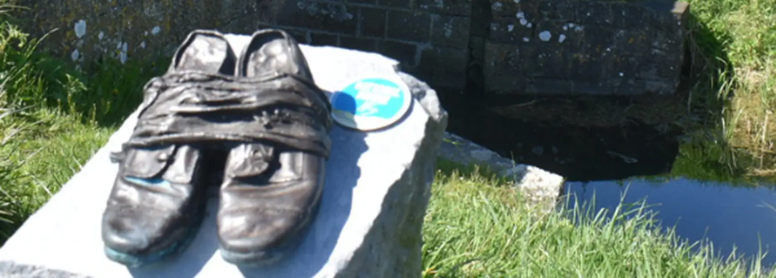 A pair of worn bronze shoes sits on a stone slab outdoors, near a small round blue plaque. In the background, there is grass and a dark water-filled stone structure.