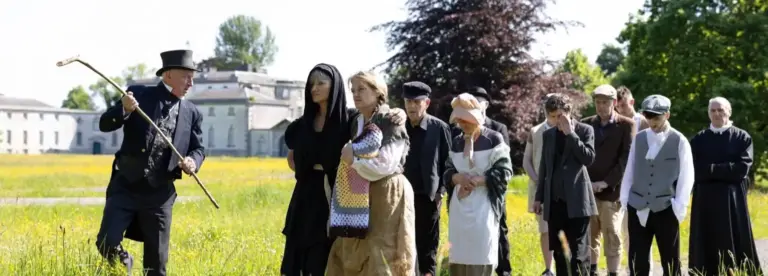 A man in an old-fashioned top hat and coat holds a walking stick, leading a queue of people in period clothing across a grassy field near a historic building, with trees and clear skies in the background.
