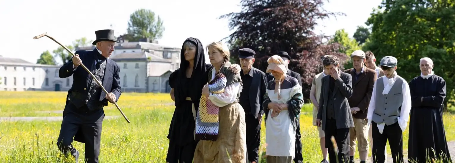 A man in an old-fashioned top hat and coat holds a walking stick, leading a queue of people in period clothing across a grassy field near a historic building, with trees and clear skies in the background.