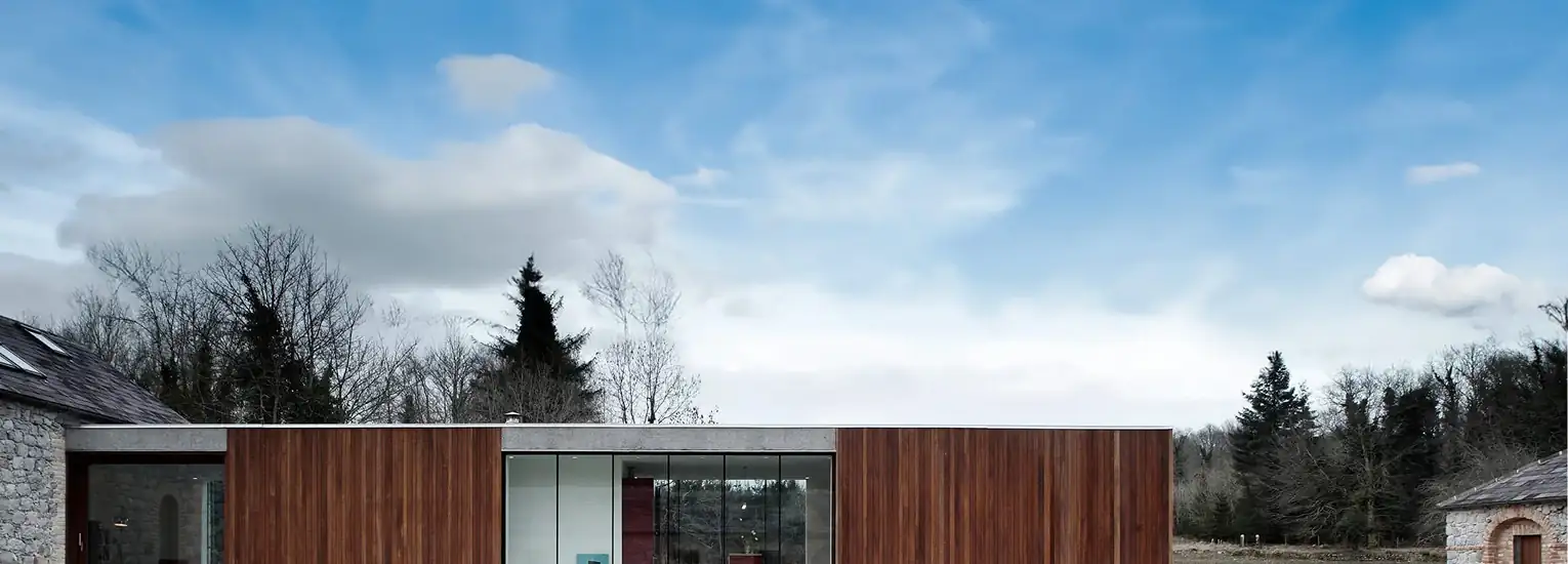 A modern, single-storey building with large glass windows and wood panelling stands in a rural landscape, surrounded by trees and grassy fields under a blue sky with scattered clouds.