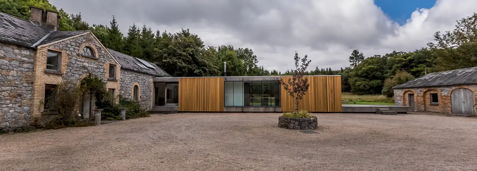 A modern building with wooden panelling and large glass windows sits between two older stone structures in a courtyard surrounded by trees under a cloudy sky.
