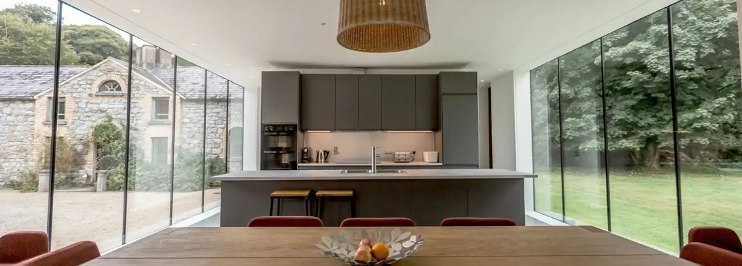 Modern kitchen with floor-to-ceiling glass walls, dark cupboards, a central island with a sink, and a wooden dining table with a fruit bowl; views of greenery and a stone house outside.