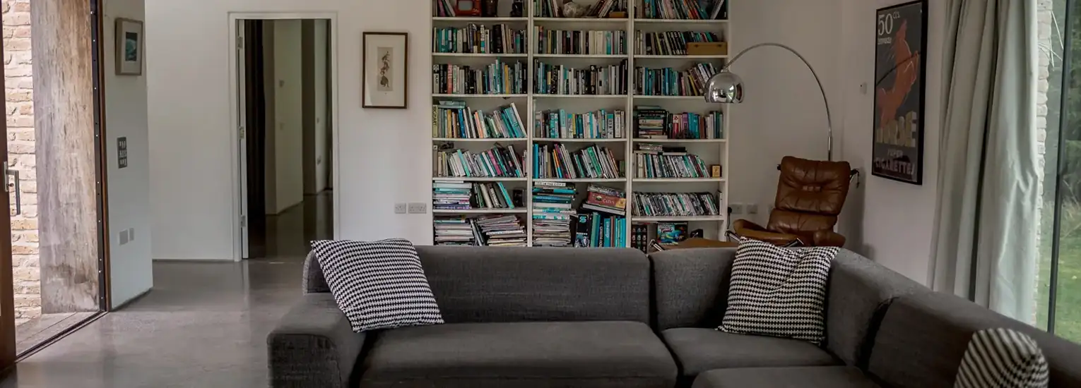 A modern living room with a grey corner sofa, striped cushions, a bookcase filled with books, a brown leather armchair, a standard lamp, a large window with curtains, and a wall poster.