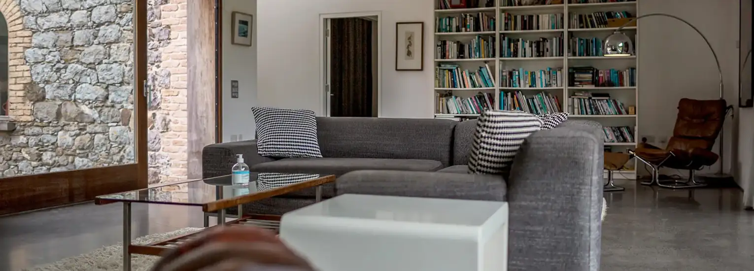 A modern living room with a grey sectional sofa, striped cushions, a glass coffee table, a bookcase filled with books, a curved floor lamp, and stone walls. Natural light enters through large windows.