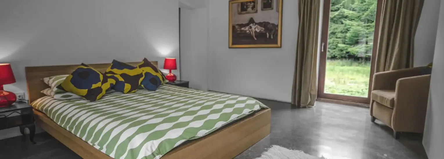 A modern bedroom with a wooden bed, green and white patterned bedding, colourful pillows, two red lamps, an armchair, a painting on the wall, and a glass door overlooking greenery outside.