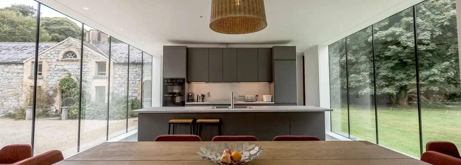 Modern kitchen with dark cupboards and an island, viewed from a wooden dining table. Large floor-to-ceiling glass walls offer views of greenery and an old stone building outside. A pendant light hangs above the island.