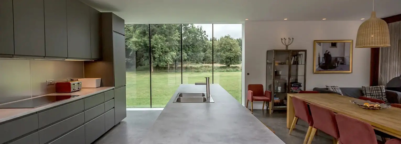 Modern kitchen and dining area with grey cupboards, a long island with a sink, red chairs around a wooden table, large glass wall overlooking a green lawn and trees, and contemporary decor.