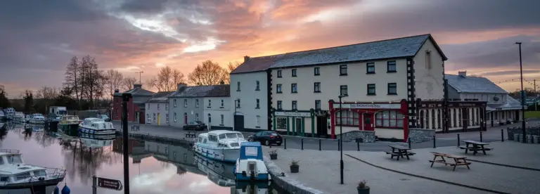 A peaceful riverside scene at sunset shows boats moored along the calm water, with a row of traditional buildings, including restaurants and shops, lining the promenade under a dramatic, colourful sky.