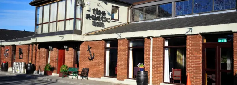 Exterior of The Rustic Inn, a brick building with large windows, outdoor benches, potted plants, barrels, and nautical decor, under a clear blue sky.