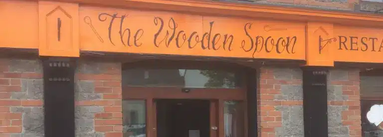 The image shows the exterior sign for "The Wooden Spoon" restaurant, with decorative wooden spoon illustrations on an orange background above a brick and stone building entrance.
