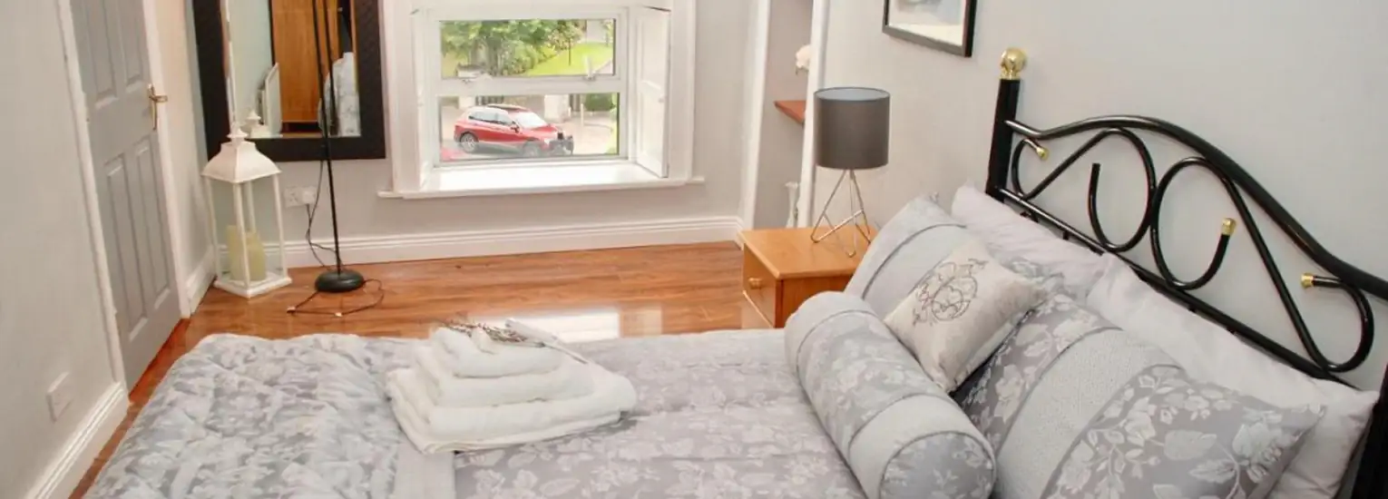 A cosy bedroom with a double bed in grey and white bedding, wooden floor, a bedside table with a lamp, a large window, a standing mirror, a lantern, and a view of a parked red car outside.